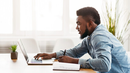 Young African American employee using laptop and taking notes while working in modern office, side view with copy space