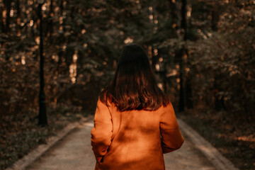 Canvas Print - Young brunette woman dressed in warm brown orange coat walking alone in autumn park. Autumnal clothes concept. Back view