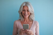 © Alexandra - Mature woman is smiling while using her phone in front of a blue background