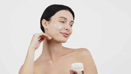 Wall Mural - A young woman cares for her clean, makeup-free skin by applying moisturizing cream to her face. The white background highlights her dedication to skincare and her desire for a healthy appearance.