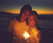 © StarDweller/peopleimages.com - Night, beach and couple with sparklers for holiday, vacation and celebrate new year together. Smile, man and woman with fireworks by sea for romantic date, travel and hug for love outdoor at sunset