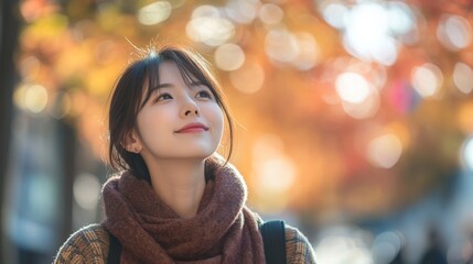 A young asian woman stands outdoors, gazing upward with a smile as vibrant autumn leaves surround her. She wears a cozy scarf, reflecting a peaceful moment in a beautiful park setting during fall.