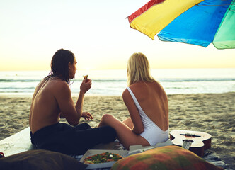 Beach picnic, summer and couple eating pizza at sunset on vacation date, relax and romantic holiday. Food, man and woman by ocean together by umbrella for travel, relationship or love with back view