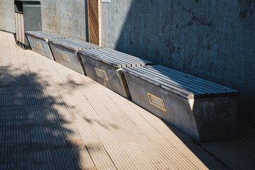 There is a row of several wooden benches placed along a sidewalk that is situated right next to a solid concrete wall, creating a comfortable space for people to sit and relax