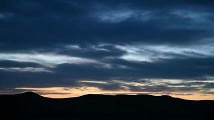 Wall Mural - Late day time lapse of blue to dark gray clouds moving above a silhouette of rolling distant hills.  The horizon is a peachy orange color.
