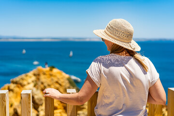 Summer vacation in Lagos Portugal. Woman in beige t-shirt, sunglasses and sun hat  standing on viewpoint on cliffs during sightseeing Ponta da piedade in Algarve on beautiful sunny day. Back view