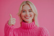 © ALL YOU NEED studio - beautiful blue eyed blonde young woman wearing knited vibrant pink sweather is smiling and showing thumb up on camera, isolated on pastel solid smooth pink background, web banner with copy space