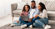 © Prostock-studio - Family Weekend. Cheerful African American parents and their little daughter using laptop at home together, watching movie or browsing internet, sitting on the floor carpet in living room
