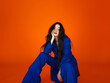 © Amy - Confident woman in bold blue pantsuit sitting against vibrant orange background, modern and trendy fashion portrait