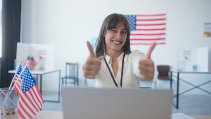 Happy girl sitting in office or polling station with American flags. Widely smiling and showing thumbs up to camera. Enjoying end of presidential elections. Motivating people to visit voting location.