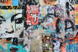 A vibrant urban wall covered in colorful graffiti and posters, featuring abstract faces and various artistic expressions. The mix of colors and textures creates a dynamic visual impact.