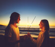 © StarDweller/peopleimages.com - Night, couple and playing with sparklers, beach and celebration for New Year, smile and bonding. Outdoor, man and woman at sea, sunset and fun for people, relationship and scenery in holiday and love