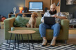 © Seventyfour - Man sitting on couch working on laptop while dog stands next to him offering company in cozy living room full of modern technology and decor including speakers, books, computer monitor