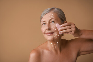 Sticker - Beautiful woman doing facial massage with gua sha tool on pale brown background. Arrows on skin showing direction of moves