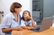 © Andrii - Granddaughter and grandmother looking at each other as the use laptop together on table. Loving senior woman and girl learn from the computer during leisure time at home