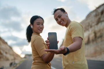 athletic runner couple wearing sportswear presenting blank screen phone, blurred background