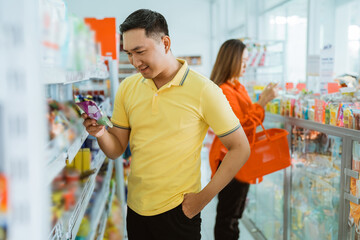 handsome man standing front of shelf while holding and looking at shopping items in supermarket