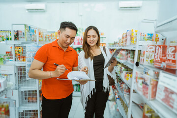 male shop assistant stands writing with clipboard with female business owner at minimarket