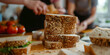 © Rohit k  - A stack of sliced whole grain bread with oats, placed on a wooden cutting board. In the background, two people are preparing food, with fresh tomatoes on a plate nearby.