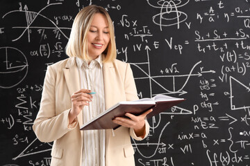 Wall Mural - Mature female math teacher with notebook and pen near blackboard in classroom