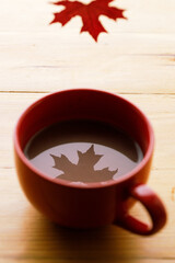 Cup of delicious hot chocolate with red maple leaf reflection on wooden table background. Selective focus.