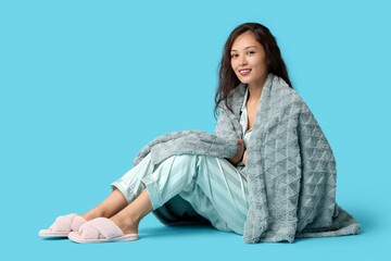 Wall Mural - Beautiful young happy Asian woman in pajamas with warm soft plaid sitting on blue background