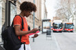 © BUDDHA - African American man in casual clothes and backpack using smartphone while standing on city street and waiting for bus