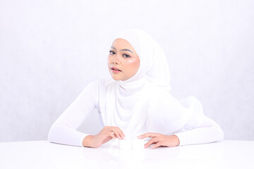 Portrait of a beautiful girl indonesia wearing elegant hijab, looking up, front view, hands holding cosmetic cream on a white table for daily facial care routine needs a white background. Perfect skin