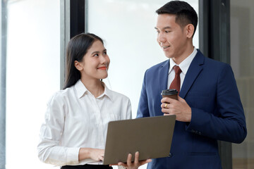 A professional woman and man engage in conversation, smiling, while holding a laptop and a coffee cup in a modern workspace.