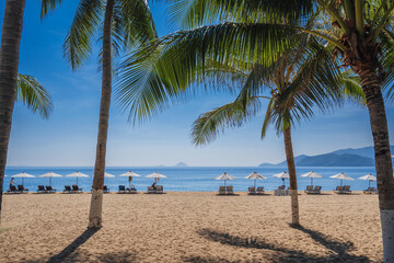 Beach umbrellas with sun beds on a sandy beach with palm trees by sea at a tropical resort in summer day in Asia