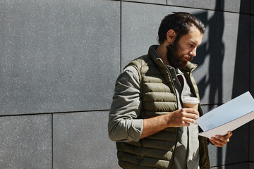 Caucasian man with beard in casual attire reading documents while holding cup in outdoor setting against tiled wall background