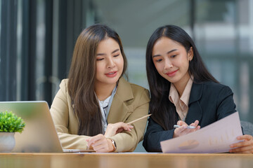 Wall Mural - Two beautiful Asian businesswomen working together using a laptop computer work together consult and advise on working together to plan work on a new product.