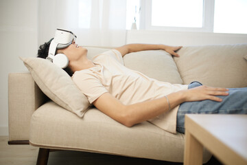 Virtual reality experience, relaxation, technology, youth, gaming lifestyle A young man wearing VR goggles lies comfortably on a sofa in a bright, modern living space