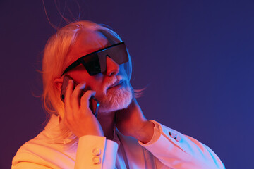 Wall Mural - Stylish man with long white hair, wearing sunglasses and a white suit, talking on the phone against a vibrant blue and pink gradient background showcasing modern aesthetics