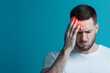 © Pongsatorn - A young man in a white shirt experiencing a headache, holding his head in pain against a bright blue background.