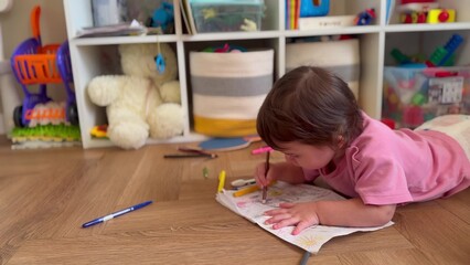 child draws in kindergarten. happy family kid dream concept. Toddler child draws at home lying on the floor. little girl in a pink jacket lies on the floor and holds colored marker close-up lifestyle