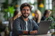 © gfx_yasir - A man with a smile, wearing a gray shirt, is sitting at a table with a laptop. Other people are in the background