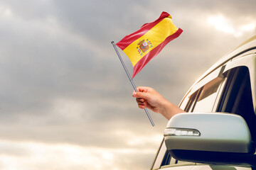 Waving the Spain flag against the sunrise or sunset from a car driving along a country road. Holding the Spain flag, traveling by car, on a weekend trip.