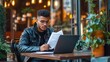 © WS Studio 1985 - A young man in glasses reads documents while working on a laptop at a cozy café surrounded by plants and warm lighting.