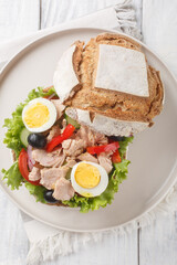 Pan bagnat is a sandwich with salad nicoise composed of raw vegetables, hard boiled eggs, anchovies and tuna closeup on the plate on the wooden table. Vertical top view from above