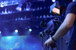 © Emvats - A videographer operates a professional camera during a live event, with vibrant blue stage lighting and blurred background creating a dynamic atmosphere. The camera screen displays the ongoing