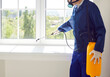 © Studio Romantic - Male exterminator from pest control service wearing protective safety mask and blue workwear suit working inside house spraying toxic liquid or gas from yellow sprayer bottle over white window sill