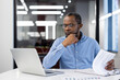 © Liubomir - Focused man in blue shirt working on financial reports at office desk. Business professional analyzing documents with laptop. Serious thinking financier accountant by paper work.
