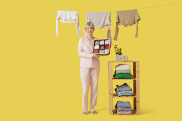 Poster - Beautiful young woman with organizer, stacks of clothes on shelf and drying laundry against yellow background