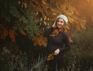 A beautiful young woman with red hair, pregnant, in brown long dress, stands in autumn park, holds her stomach with one hand, hair  blowing in the wind. foreground the branche maple with yellow leaves