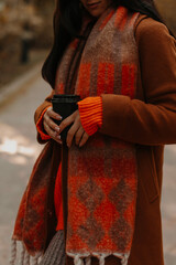 Poster - Woman dressed in warm brown coat, knitted sweater and orange scarf drinking coffee on the street, concept of cozy autumn walk