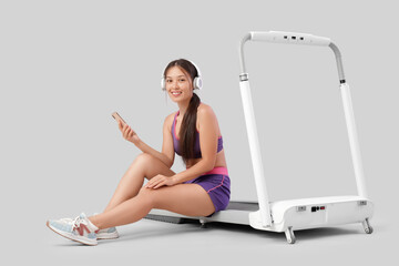 Canvas Print - Sporty Asian woman in headphones using mobile phone on treadmill against light background