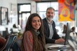 © Joaquin Corbalan - A young Latina woman in office attire smiles while working at a computer, with a colleague engaged in tasks at a modern workspace bustling with activity