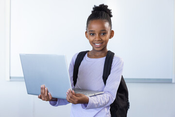 Wall Mural - In school classroom, african american girl holding laptop and smiling with backpack