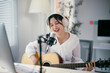 © Parichat - Young woman is recording music at home, playing her guitar and singing into a microphone. She is wearing headphones and using a computer to record her music